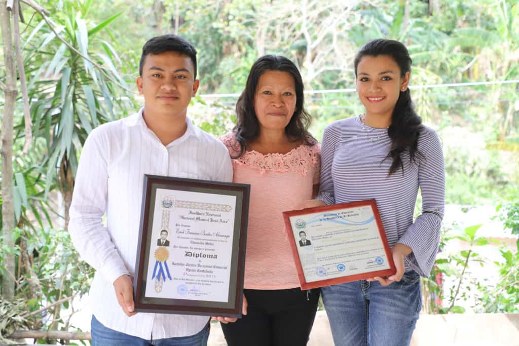 Erick, wearing a white striped shirt and jeans, is standing with his mother, Teresa, wearing a pink shirt and black pants, and his sister, wearing jeans and a purple shirt. Erick and his sister are holding Erick's diplomas. There are trees in the background.