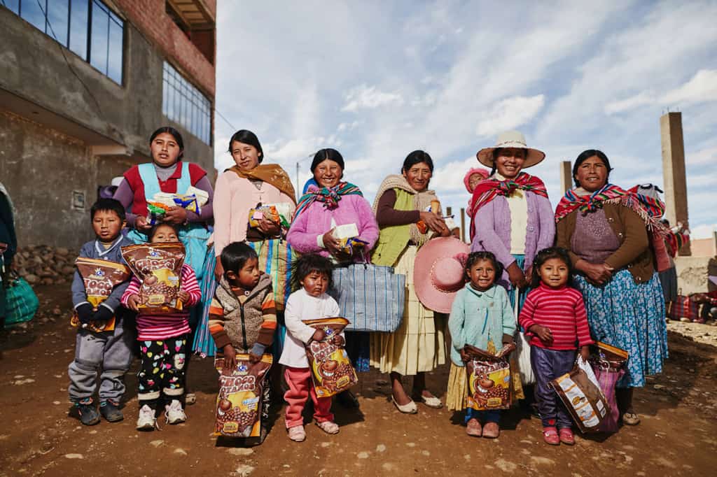 A group of mothers wearing blue, pink, green, purple and brown sweaters, and children stand on a dirt road holding bags of food. They are posing for a picture next to a building.