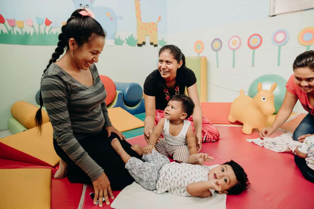 Mothers and young children playing on a red mat in a brightly colored play room.