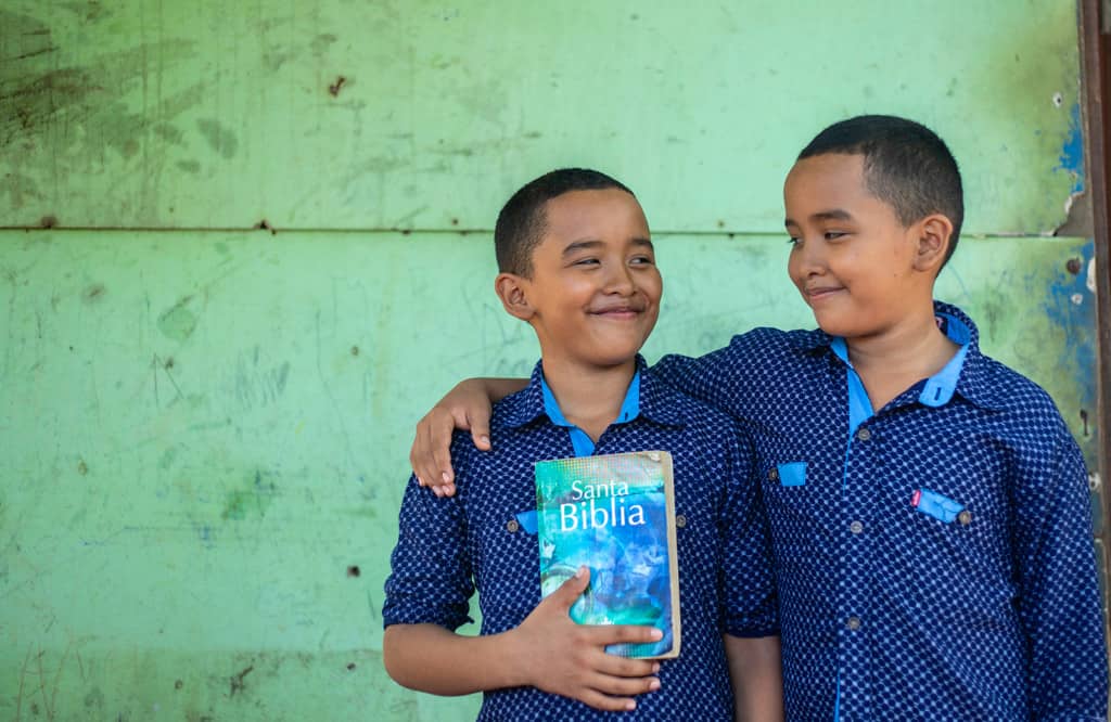 Twin boys wearing blue shirts and black pants. They are standing in front of their home and one is holding a Bible. The other has his arm around his brother.