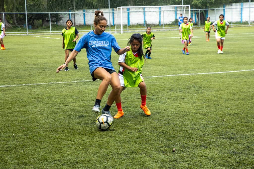 A woman in a blue Team Up with Compassion shirt is playing soccer with a ball on a field with girls in red and white uniforms covered with green pinnies.