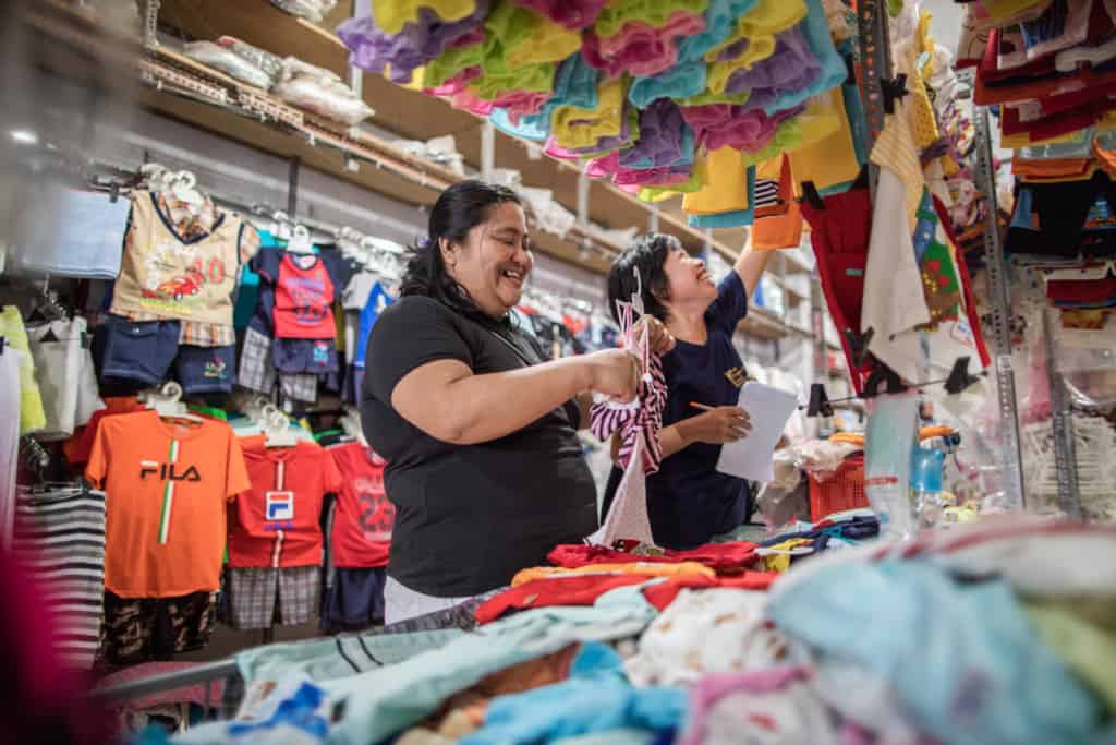 Two women are smiling and laughing as they are choosing items in the store. They are surrounded by clothing.