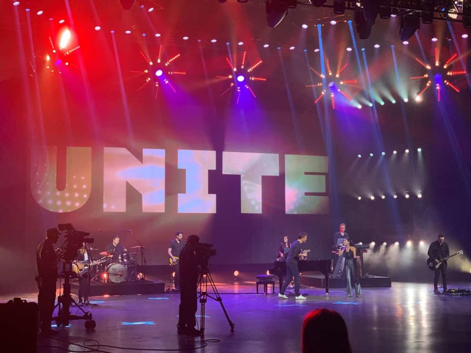 an image of the Unite to Fight Poverty concert shows band members and camera people on a large stage with bright red and purple lights all around them and the word Unite on the wall behind the stage