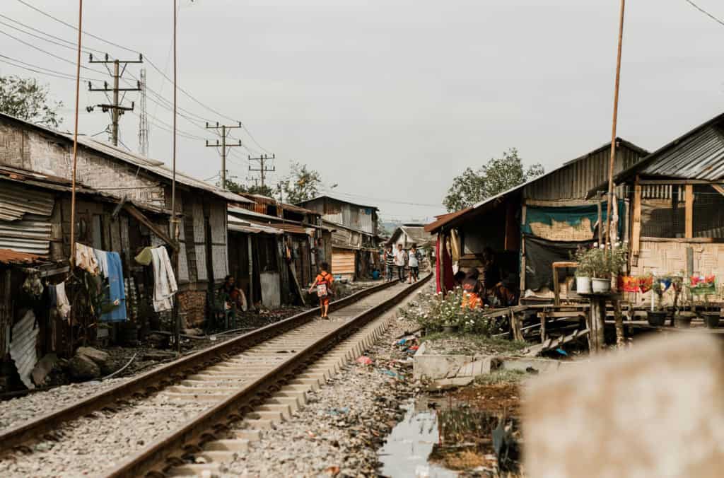 View of a child, wearing a backpack and orange shirt, walking away down a train track with extreme living conditions, impoverished homes lining the tracks in a slum neighborhood.