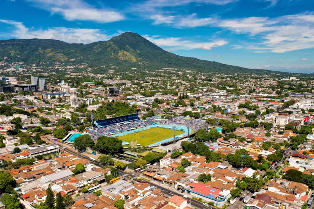 Urban aerial shot of a city with a stadium. There are buildings surrounding the stadium and a green mountain in the background.