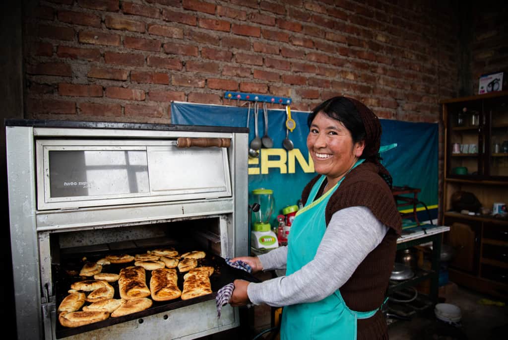 Woman wearing a brown and gray shirt and a green apron. She is taking a pan of her baked goods out of the oven. The background is a brick wall.