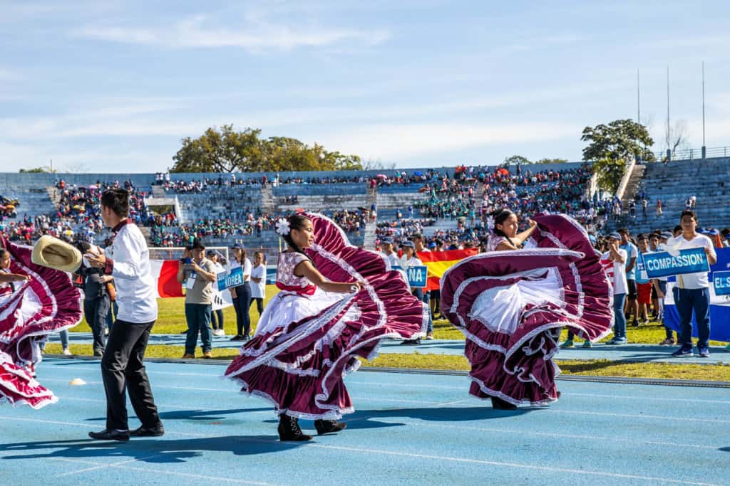 Young women in red and white dresses are dancing on the track inside the stadium. There are people on the grass holding signs and people sitting in the stands in the background.