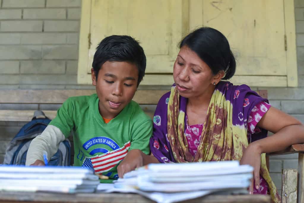 a boy wearing a green shirt and his mother, wearing a colorful purple and yellow outfit, look over school papers, homework.