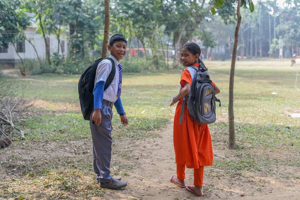 a boy and girl, brother and sister, walk down a dirt path wearing backpacks, heading for school