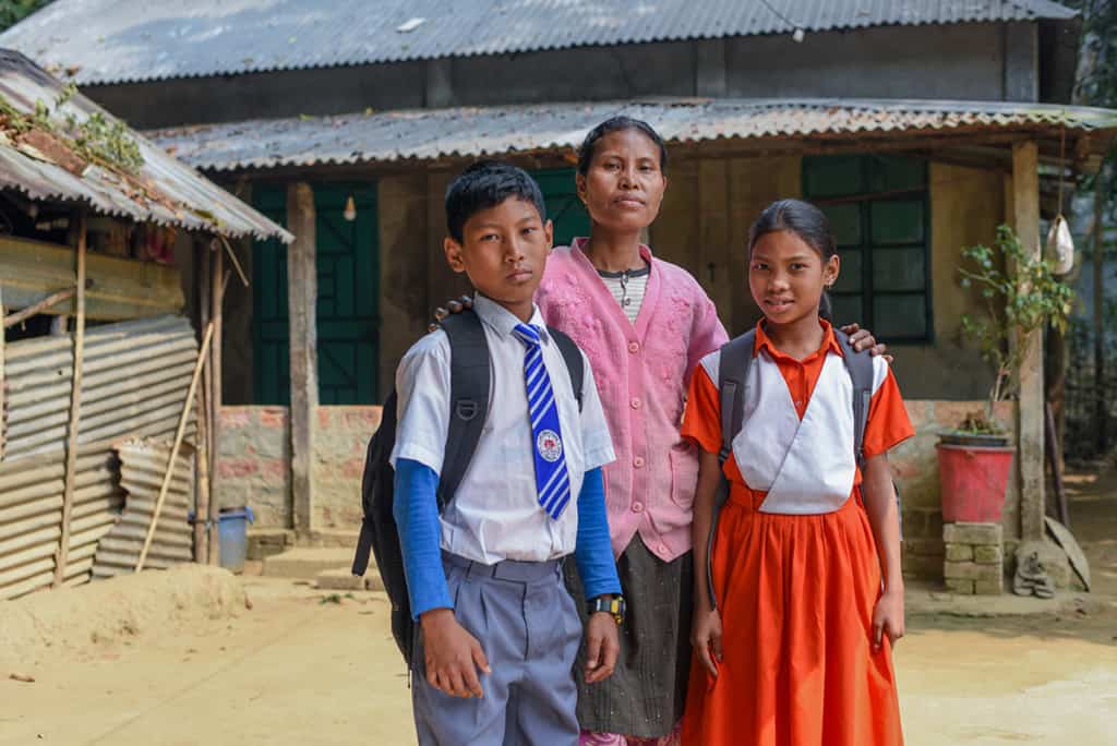 a mother wearing a pink shirt stands behind a boy and girl, each wearing a backpack. The boy wears a white shirt and blue tie. the girl wears an orange and white dress