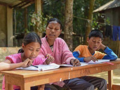 A woman, a girl and a boy sit at a long, narrow desk outdoors. The woman and girl are looking over papers, schoolwork. The boy is concentrating on his papers, homework.