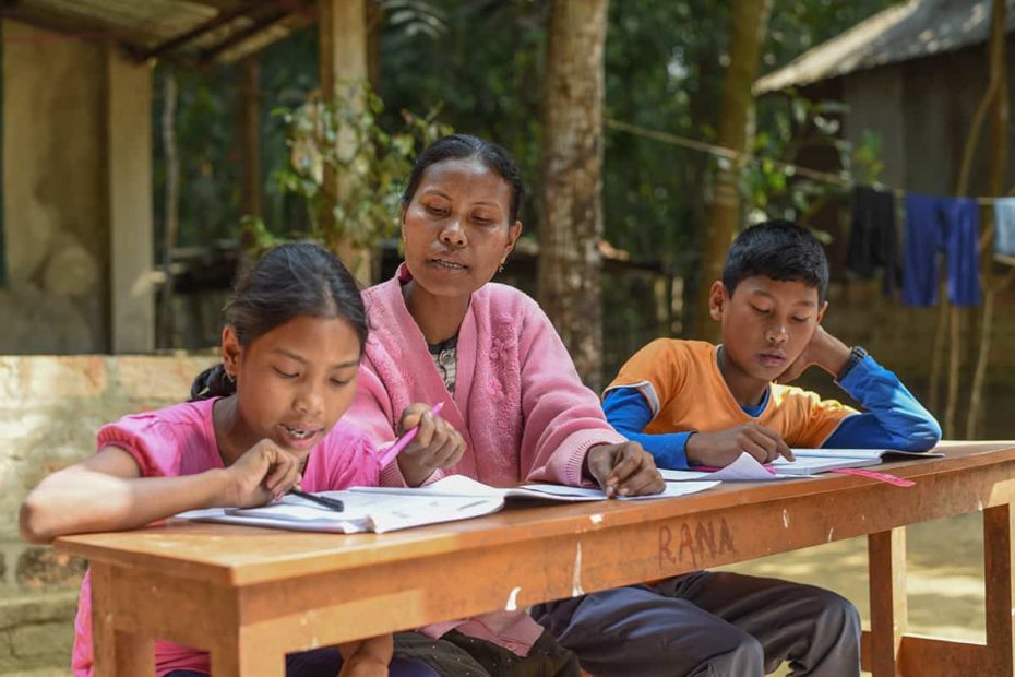 A woman, a girl and a boy sit at a long, narrow desk outdoors. The woman and girl are looking over papers, schoolwork. The boy is concentrating on his papers, homework.