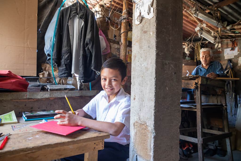 A boy wearing a white shirt sits at a wooden desk in his home school. He has school supplies in front of him on the table. A man works at a separate desk in the background.
