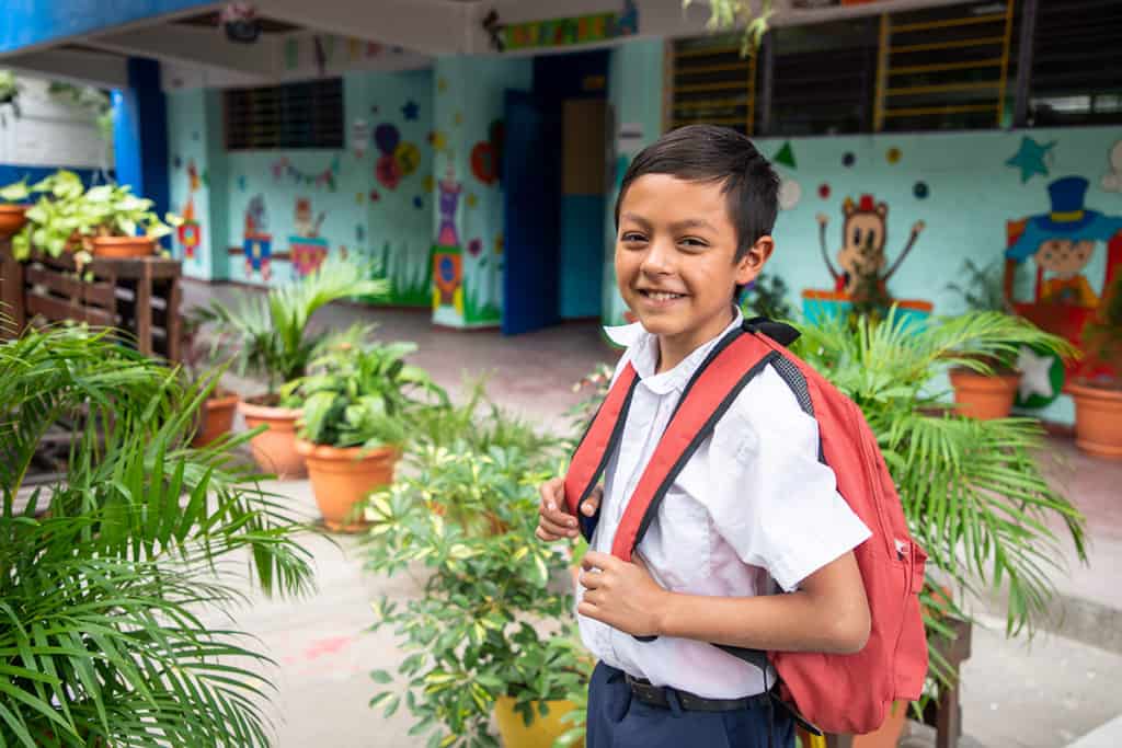 A boy wearing a white shirt and red-orange backpack smiles at the camera and looks ready for school. He is outside of a brightly colored building with many potted plants around him.