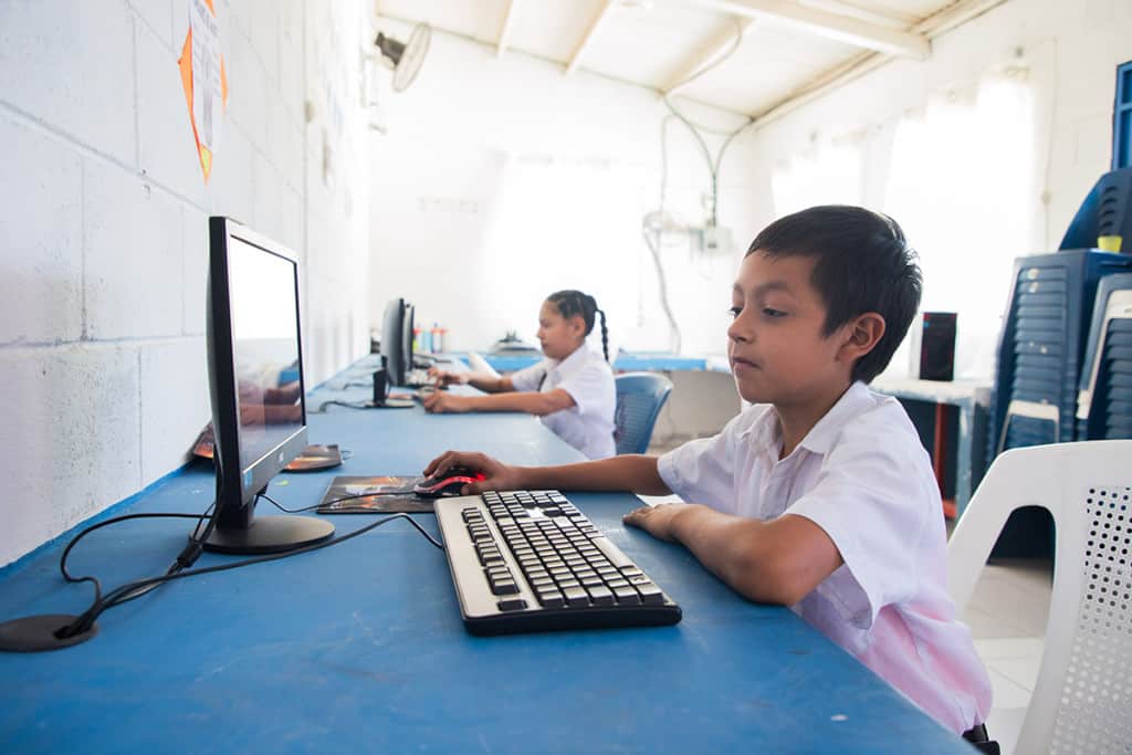 A young boy wearing a white shirt works on a computer. Another child works on a computer nearby. The computers are on a blue table.