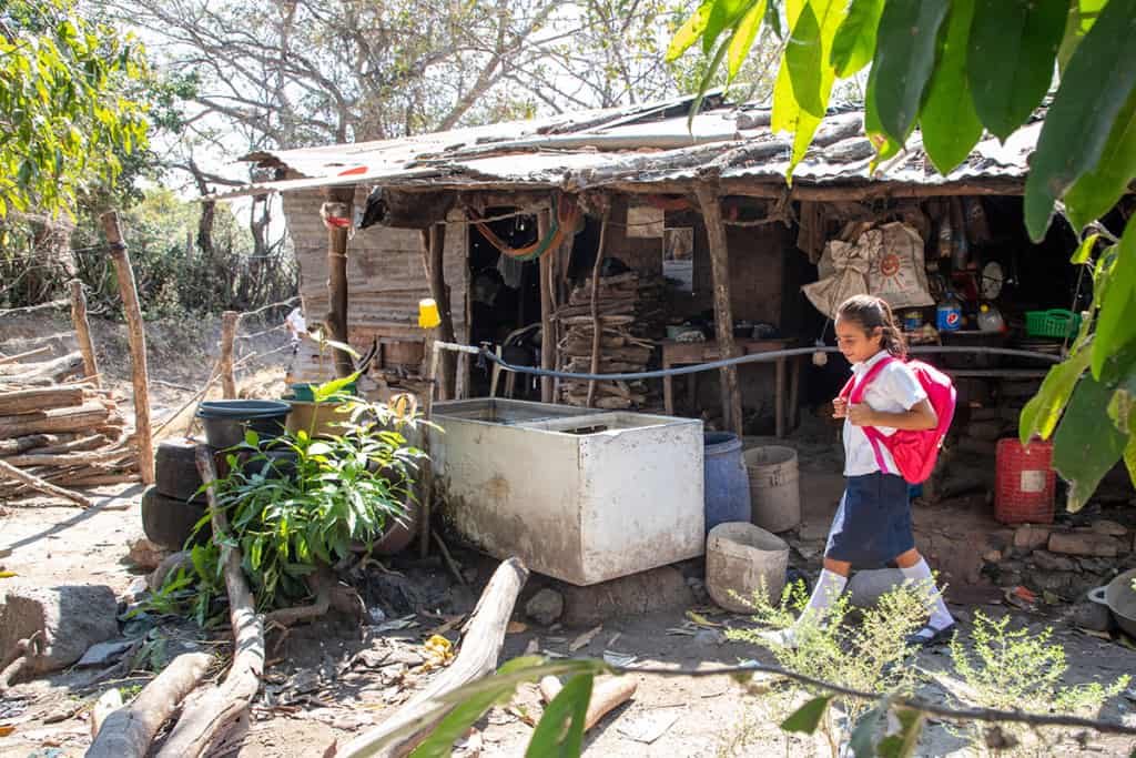 a girl wearing a white shirt, blue skirt and red backpack walks past a small wooden home.