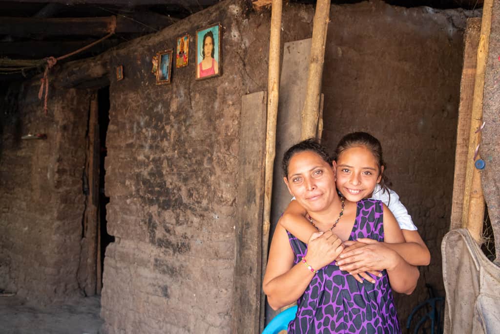 A girl stands behind a woman wearing a purple shirt and hugs her.