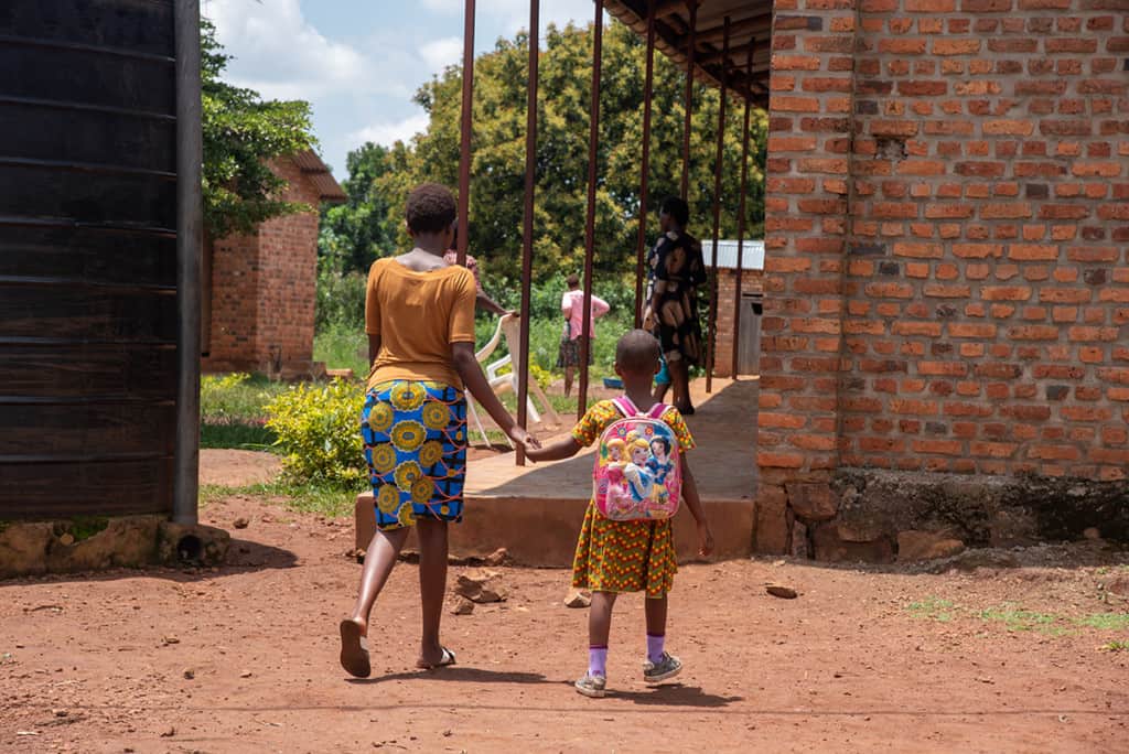 a mother and daughter dressed in brightly colored clothing walk hand in hand on a dirt road.