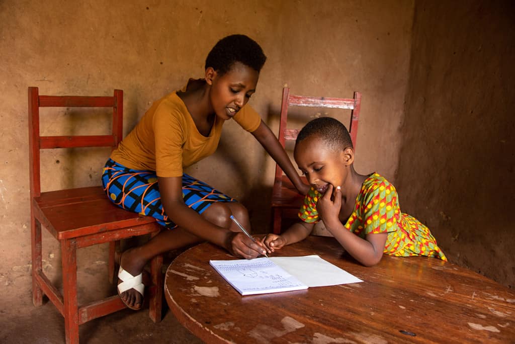 A woman and a young girl lean over papers on a wooden table. The mother is sitting on a wooden chair, and the girl concentrates on the papers, home school work.