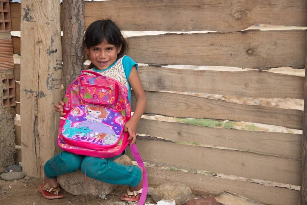 A 5-year-old girl with a shy expression holds a large pink backpack. She is wearing teal and white clothing. A wooden wall is behind her.