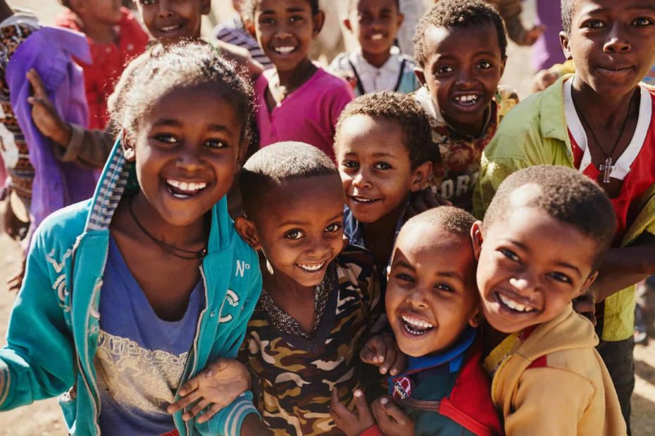 A group of smiling and laughing children wearing blue, camouflage, green, red and yellow are crowded together. There are more children in the background.