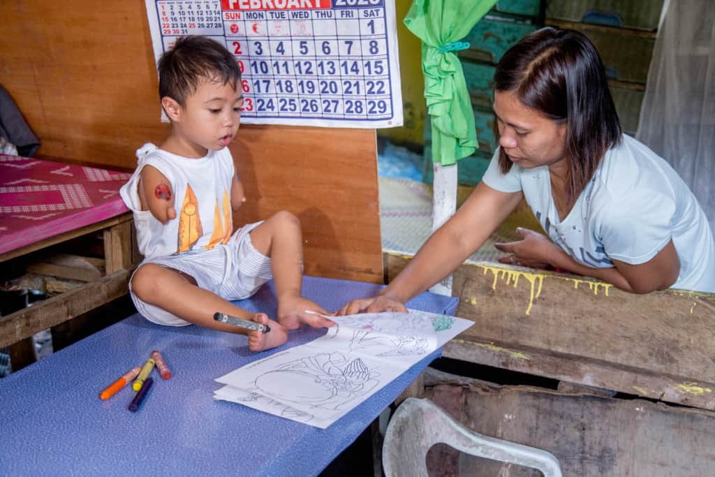 Jasper is sitting on a blue table using his feet to color in a coloring book. His mother, wearing a white shirt, is standing beside him. Jasper is wearing a white shirt and gray and white striped shorts. There is a calendar on the wall behind him.