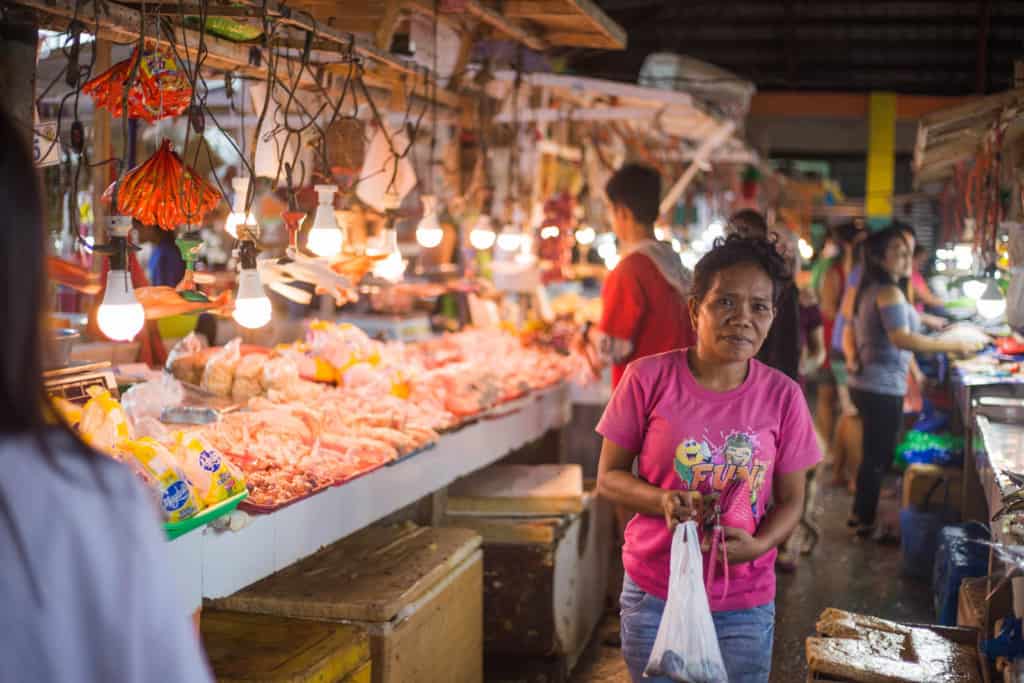 Woman wearing a pink shirt and is at the market near a milkfish stand. There are lights above the table.