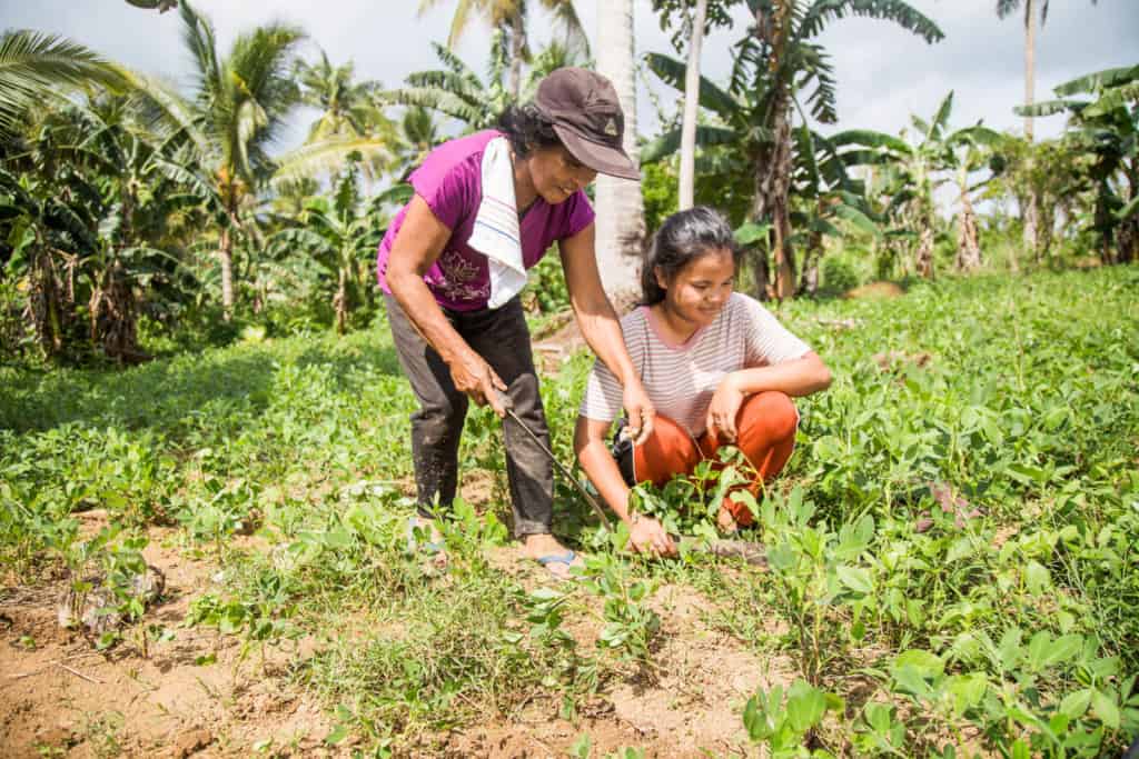 Joan and her mother, Marlyn, are working together in a field. Joan is wearing red pants and a pink and white striped shirt. Marlyn is wearing a purple shirt and gray pants.