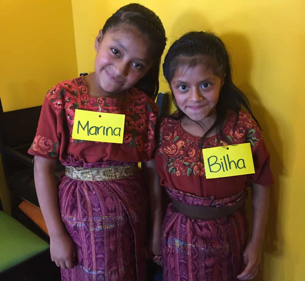 Two young girls, sisters, wearing traditional Guatemalan clothing of skirts and floral blouses. They are smiling.