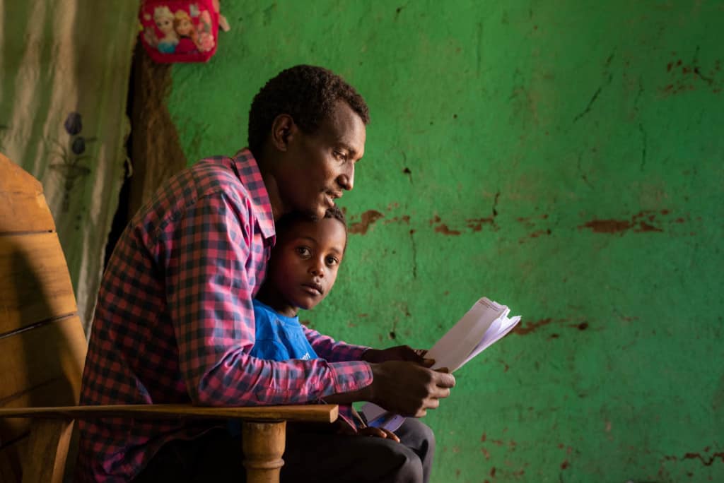 Kenenisa is wearing a blue shirt. He is sitting on his father's lap while his father reads his sponsor's letter to him. Kedir is wearing a red and black checked shirt. The wall behind them is green.