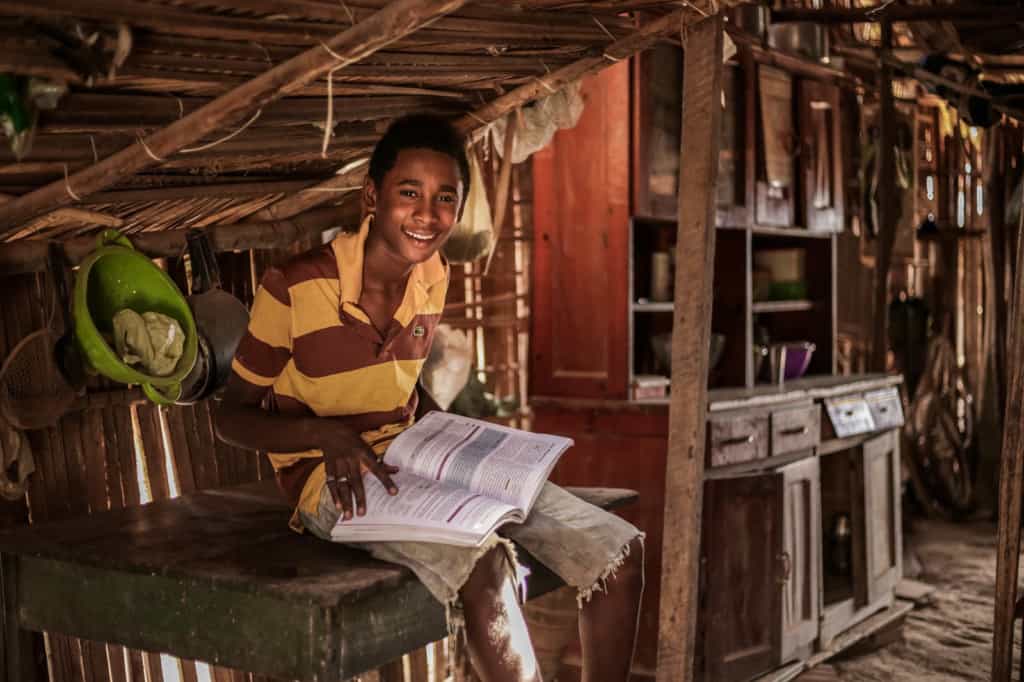 João is sitting on his kitchen table and is reading a book. He is wearing a brown and yellow shirt.