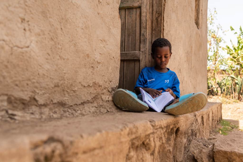 Kenenisa is wearing a blue shirt. He is sitting on a small ledge by the front door of his house and is looking through his sponsor's letters. His house is tan.