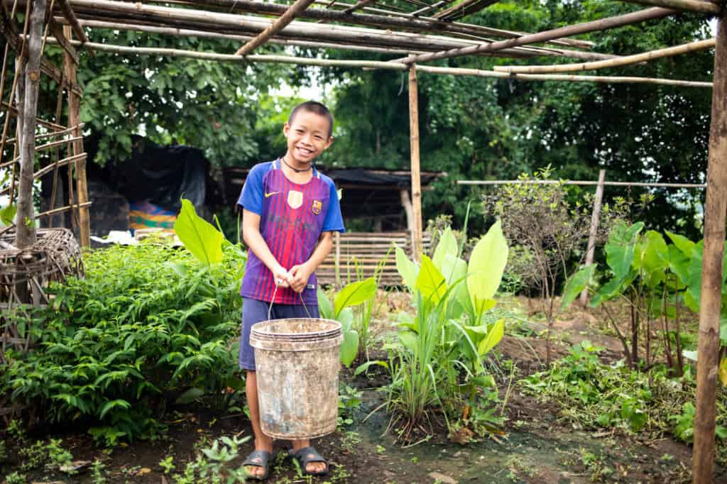 Boy wearing a blue and maroon shirt with blue shorts. He is standing outside his home and is holding a bucket full of food for his pigs. There are plants surrounding him.