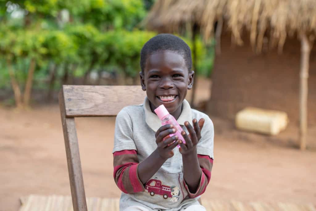 Boy weariung a gray shirt and is sitting in a chair outside his home. He is holding up a bottle of hand sanitizer.