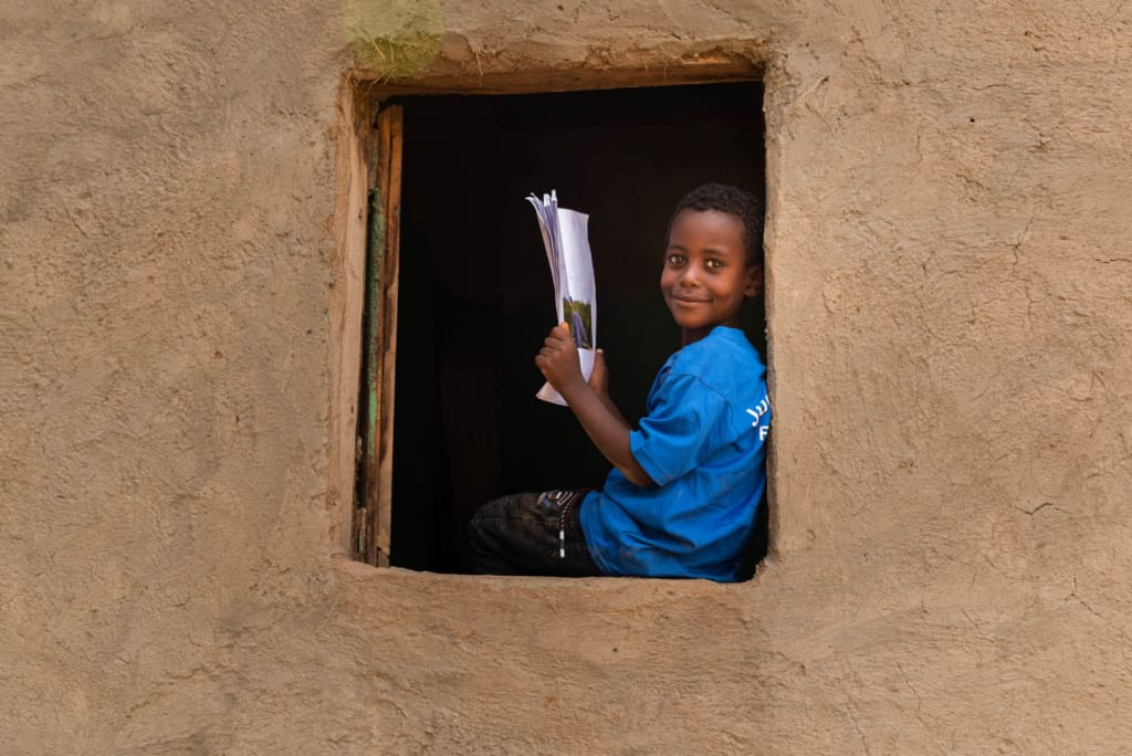 Kenenisa is wearing a blue shirt. He is sitting in an open window at his home and is holding up his sponsor's letters. His home is tan.