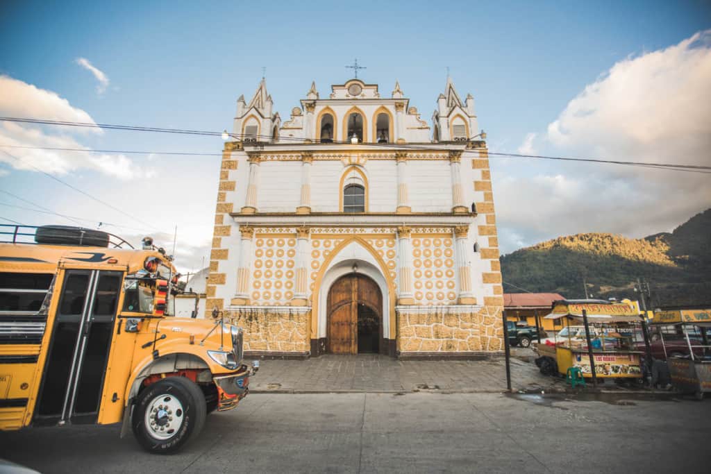 A yellow and black school bus is parked in front of a gold and white church building with blue sky and clouds in the background.