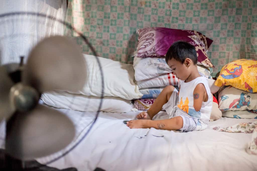 Jasper is sitting on his bed at home and he is using a cell phone with his feet. In the foreground of the picture is a fan. Behind his is a blue and white sheet hanging up as a room separator. Jasper is wearing a white shirt and gray and white striped shorts.