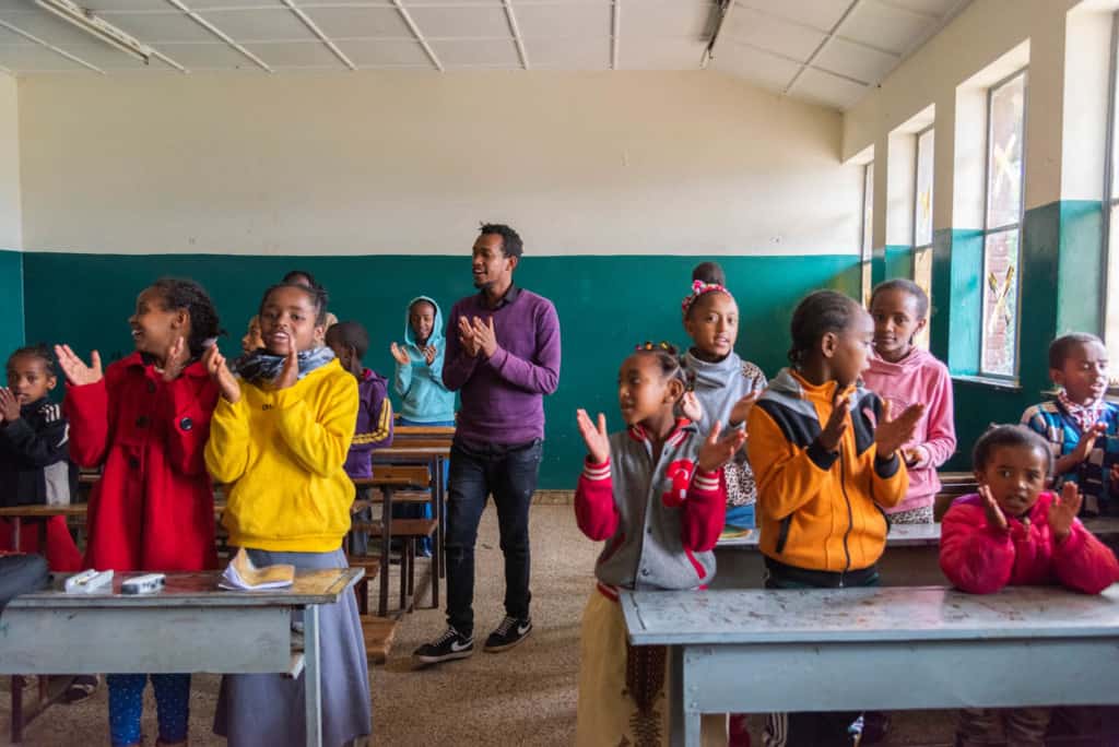 Teacher wearing a purple sweater and black pants. He is standing in a Compassion classroom with a group of children. They are all singing and clapping their hands together.