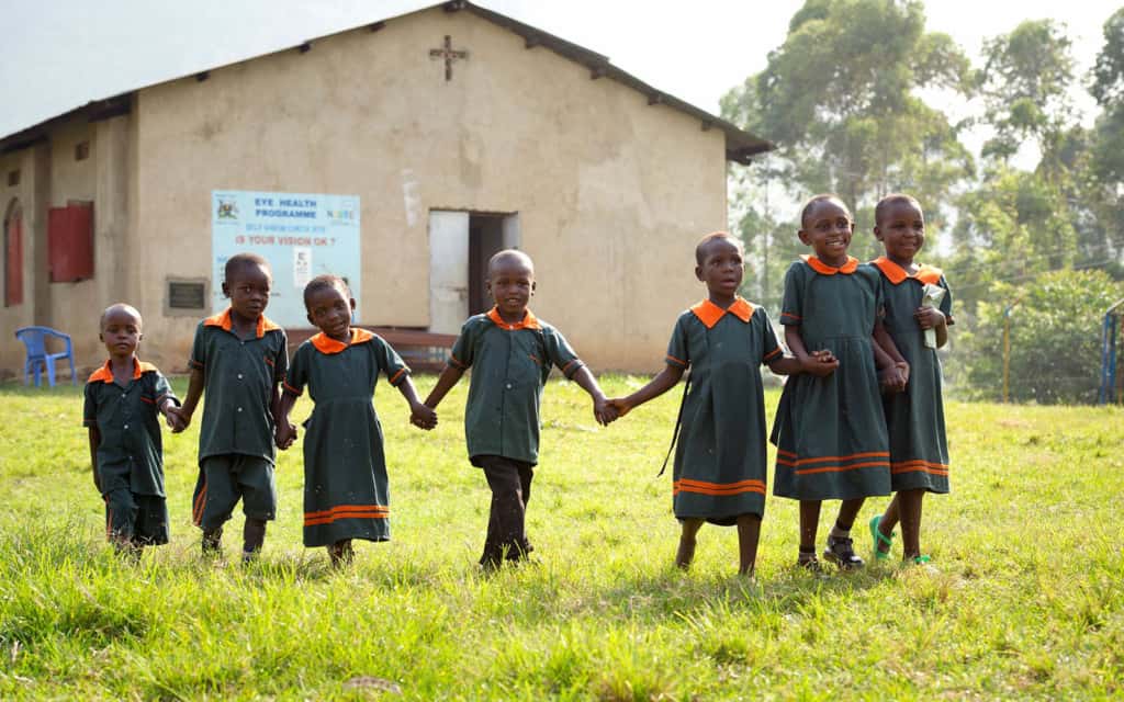 Children in uniforms holding hands in from a of church. They are standing on grass.