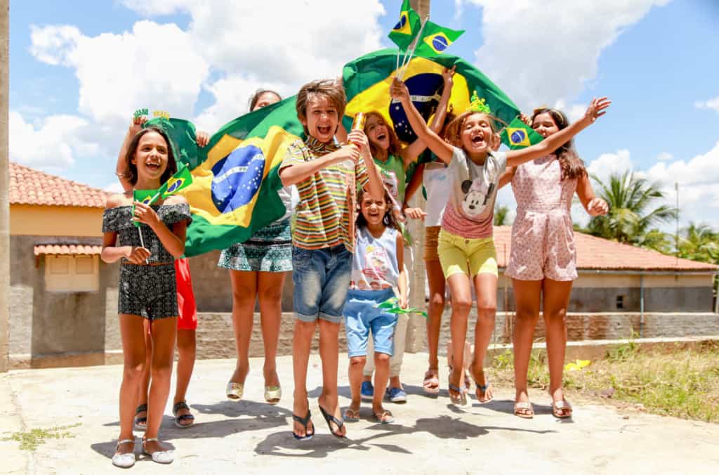 A group of children in Jericó, Brazil, cheer and laugh holding Brazilian flags. Some children are jumping, dancing.