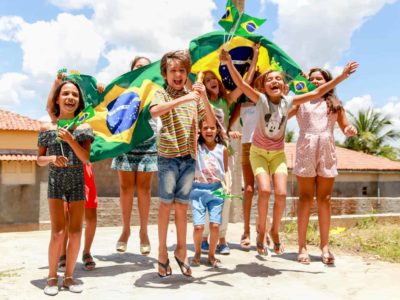 A group of children in Jericó, Brazil, cheer and laugh holding Brazilian flags. Some children are jumping, dancing.