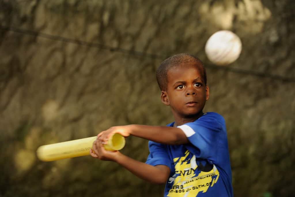 Boy swings a bat outside in his yard near his home, keeping his eyes concentrated on the white ball. He is wearing a blue shirt.