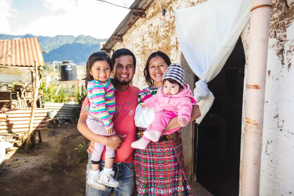 A family, mom, dad and two siblings pose for a picture outside the doorway of a white and brown house with a white curtain, fabric hanging in the door opening. The baby wears a pink sweatshirt and pants, the other girl wears a pink, blue and yellow shirt. There are hills in the distance.