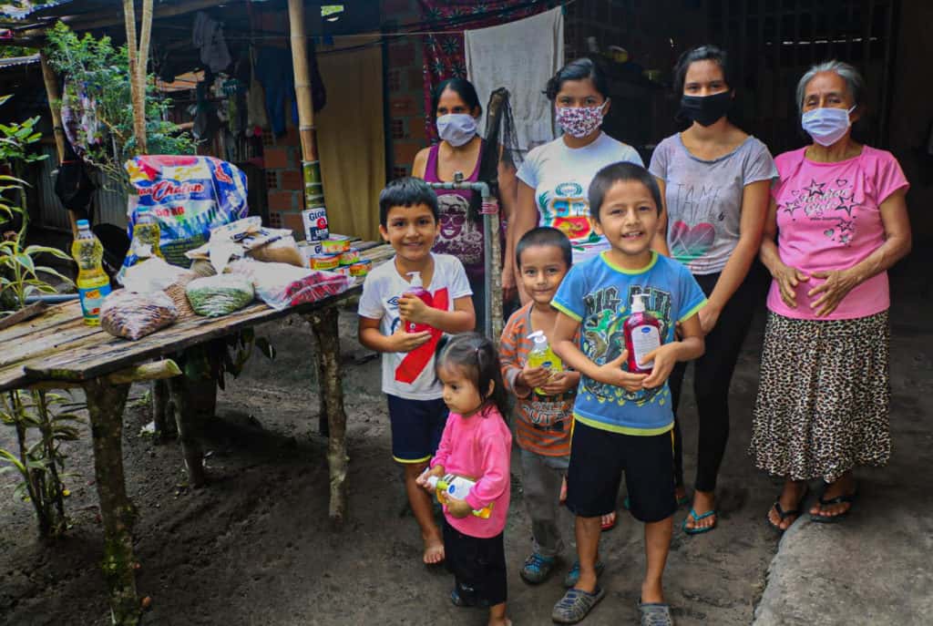 Children standing and holding some supplies. Adults are standing behind the boys and next to the food brought by the Compassion center. The girls are all wearing face masks. They are all outside their home.