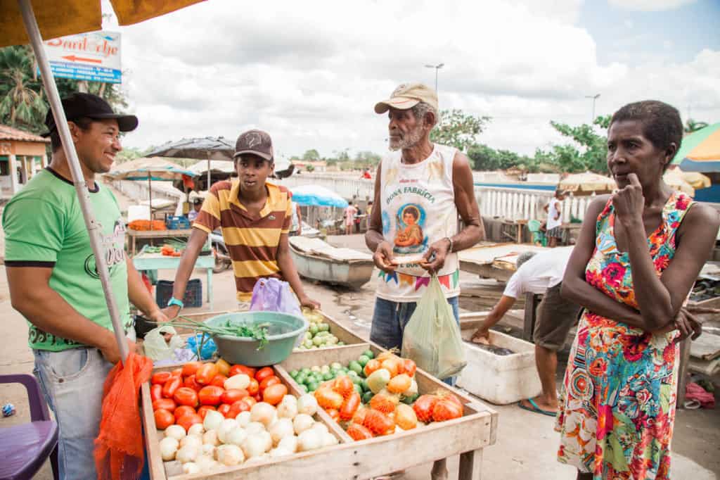 João, wearing a yellow and brown shirt, is at the market with his father, wearing a white shirt with a design on the front. They are buying vegetables.