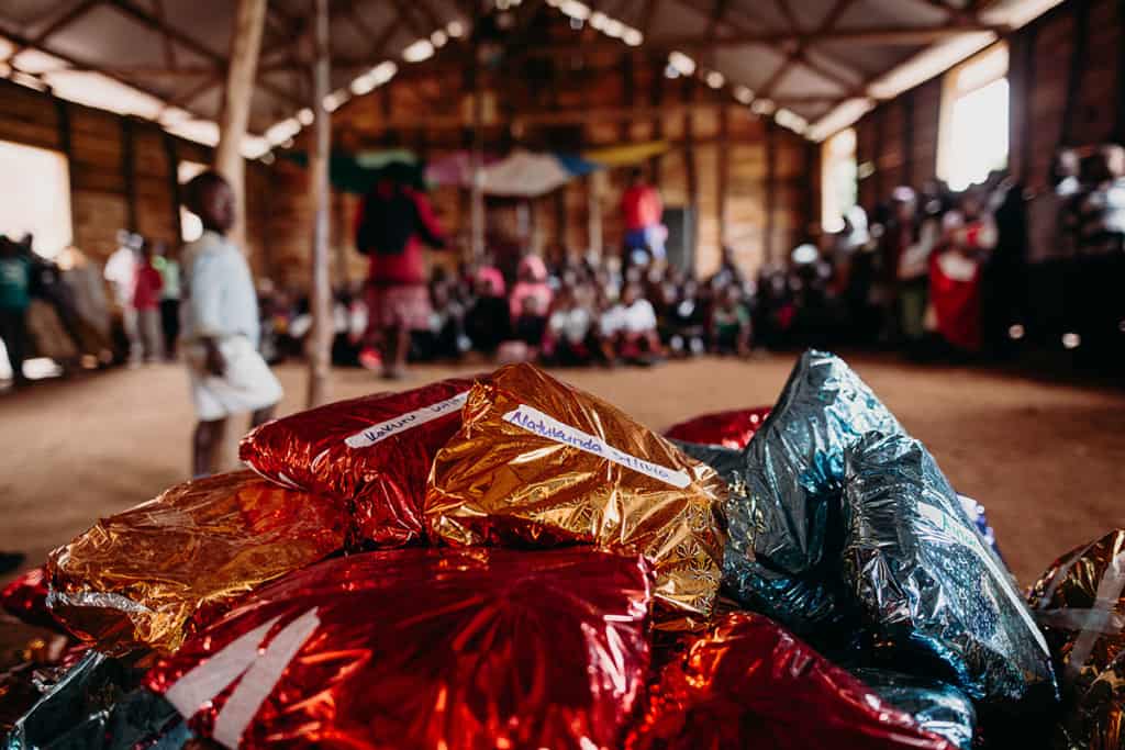 a pile of presents in shiny wrapping paper is seen in the foreground, while children sit in the background inside the barnhouse church