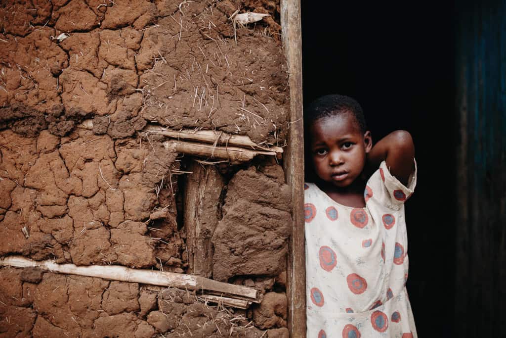Girl looking at the camera and standing in the doorway of a mud building. She is wearing a white dress with large red polka dots.