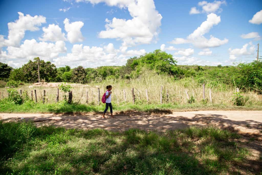 Estefany is walking down a dirt raod on her way to school. She is wearing jeans and a pink backpack. There are trees on either side of the road.