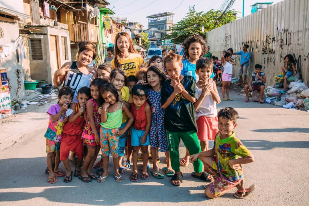 Group of children, posing for a photo. There are buildings and more people along a tall wall, fence in the background.