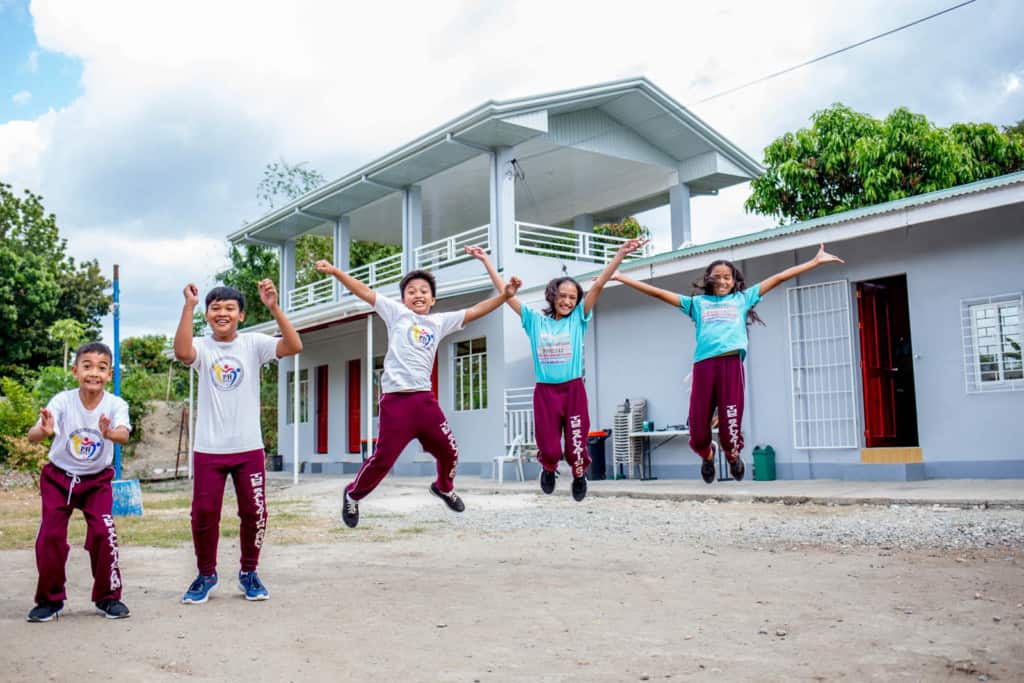 Children jumping and laughing in front of the child development center buildings, which are white with red doors. The boys are wearing white uniform shirts and the girls are wearing blue, turquoise.